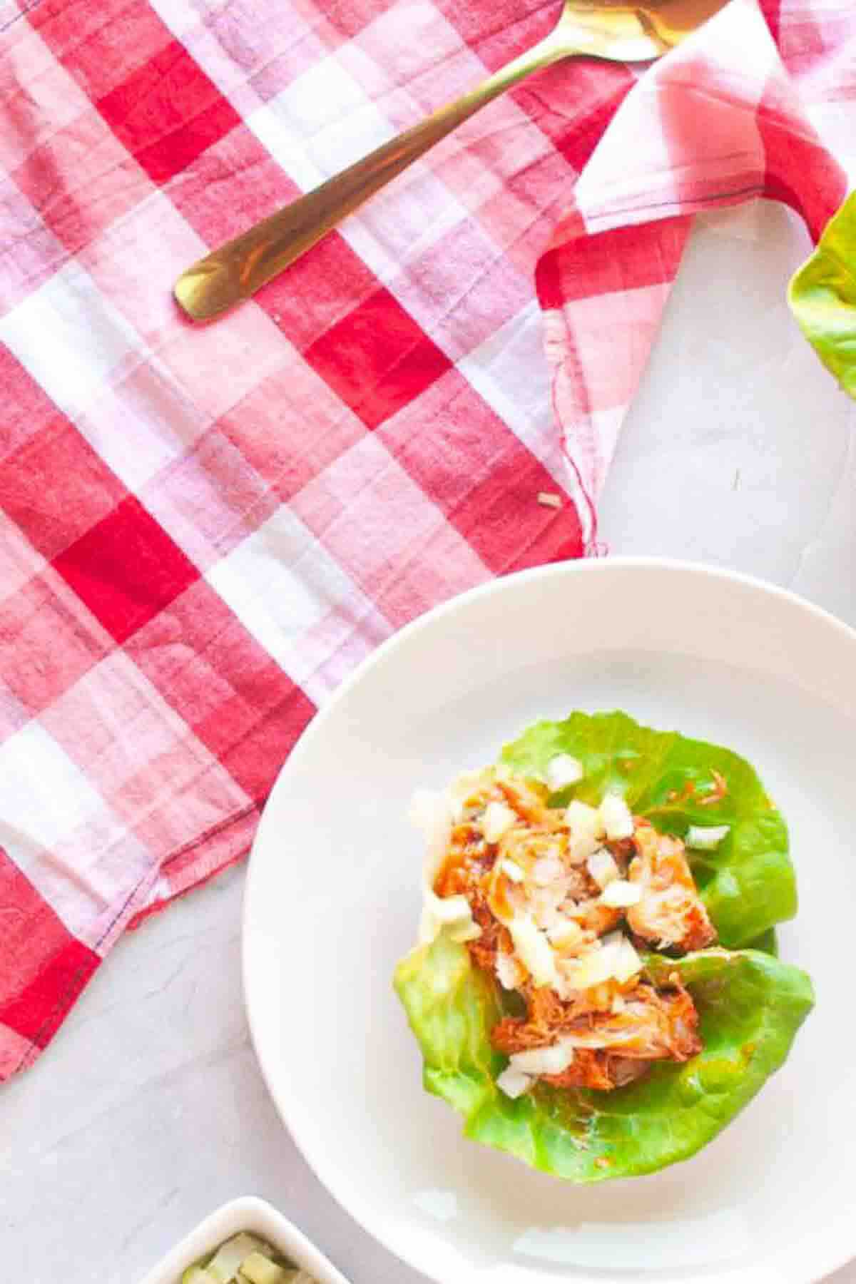 A lettuce leaf topped with shredded meat and diced onions on a white plate, next to a red and white checkered cloth and a gold spoon.
