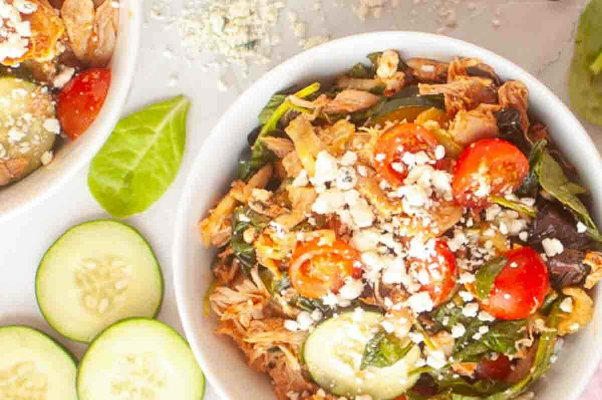 A bowl of shredded chicken salad with cherry tomatoes, cucumber slices, mixed greens, and crumbled cheese, with cucumber slices and spinach leaves nearby.