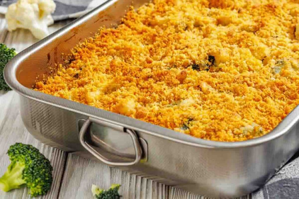 A metal baking dish filled with baked casserole topped with a golden breadcrumb crust; pieces of broccoli and cauliflower are nearby on the table.