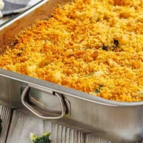 A metal baking dish filled with baked casserole topped with a golden breadcrumb crust; pieces of broccoli and cauliflower are nearby on the table.