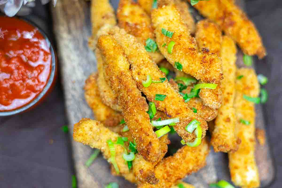 Breaded and fried chicken strips garnished with chopped green onions, served on a wooden board with a side of red dipping sauce.