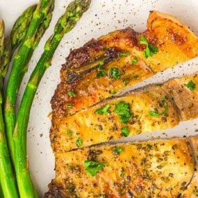 Grilled asparagus spears and sliced, seasoned chicken breast on a white plate, garnished with chopped herbs and black pepper.