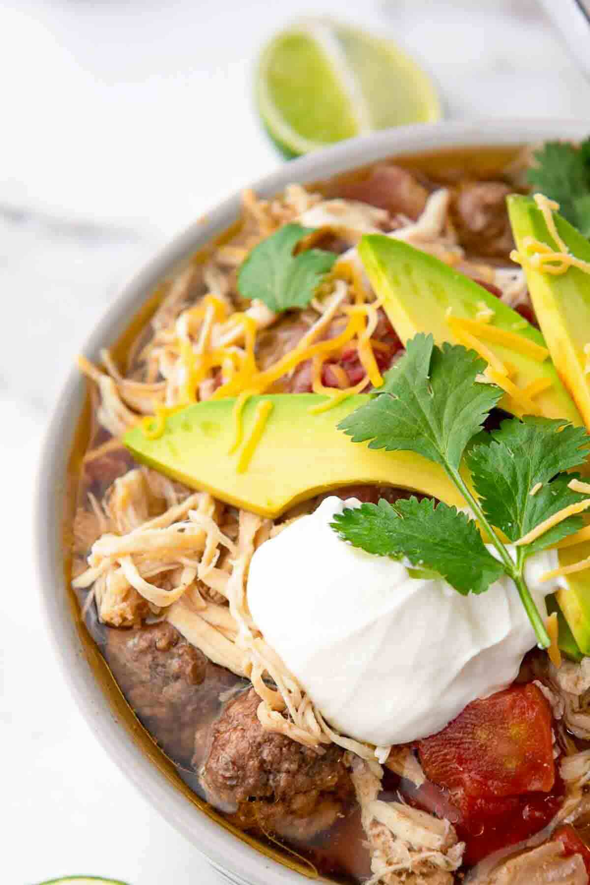 A bowl of soup topped with shredded chicken, sliced avocado, shredded cheese, sour cream, cilantro, and tomato pieces, with a lime wedge in the background.