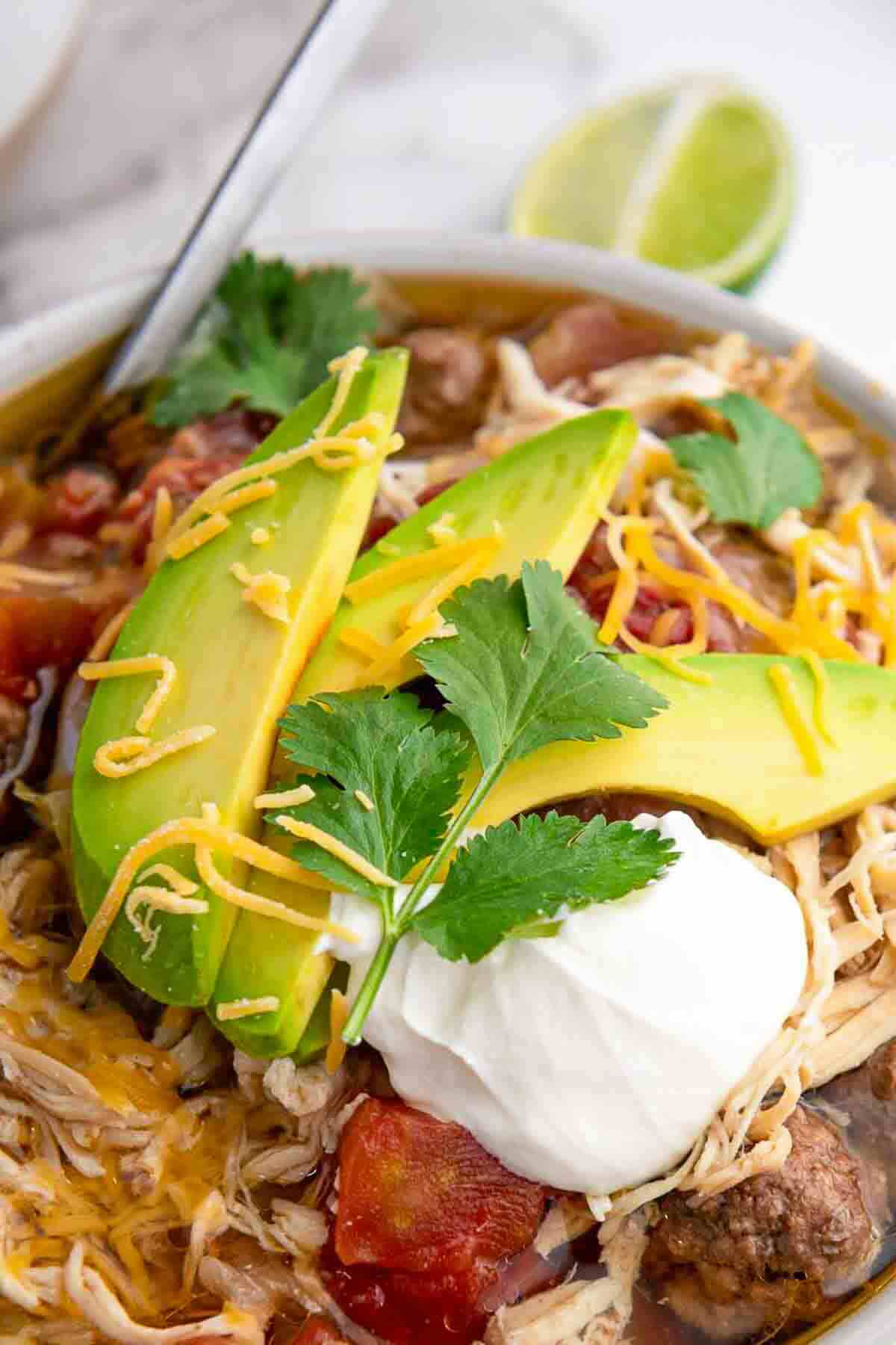 A bowl of shredded chicken soup topped with avocado slices, shredded cheese, sour cream, fresh cilantro, and tomato chunks, with a lime wedge in the background.