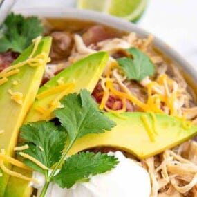 A bowl of shredded taco soup topped with sliced avocado, shredded cheese, cilantro, and a dollop of sour cream.