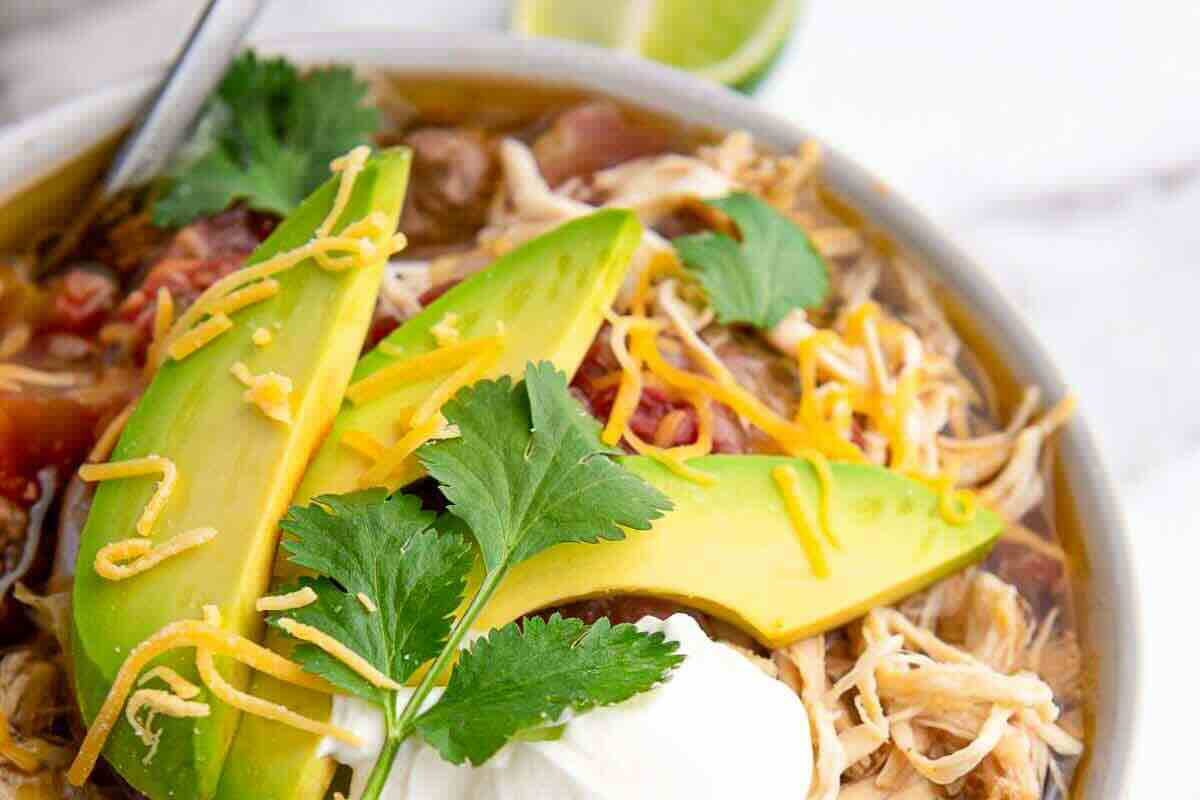 A bowl of shredded taco soup topped with sliced avocado, shredded cheese, cilantro, and a dollop of sour cream.