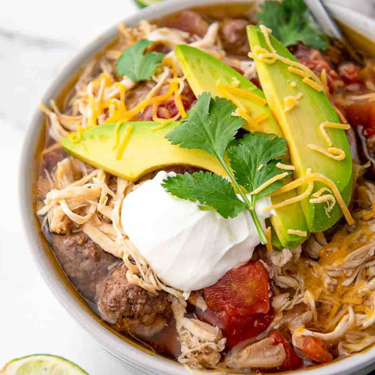 A bowl of shredded chicken and beef chili topped with avocado slices, shredded cheese, sour cream, and cilantro.