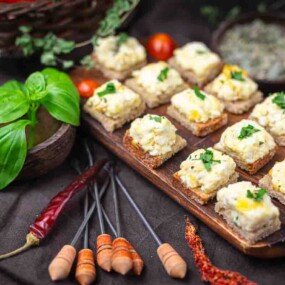 A wooden board displays several pieces of bread topped with a creamy mixture and herbs, surrounded by fresh basil, tomatoes, dried chili peppers, and skewers.