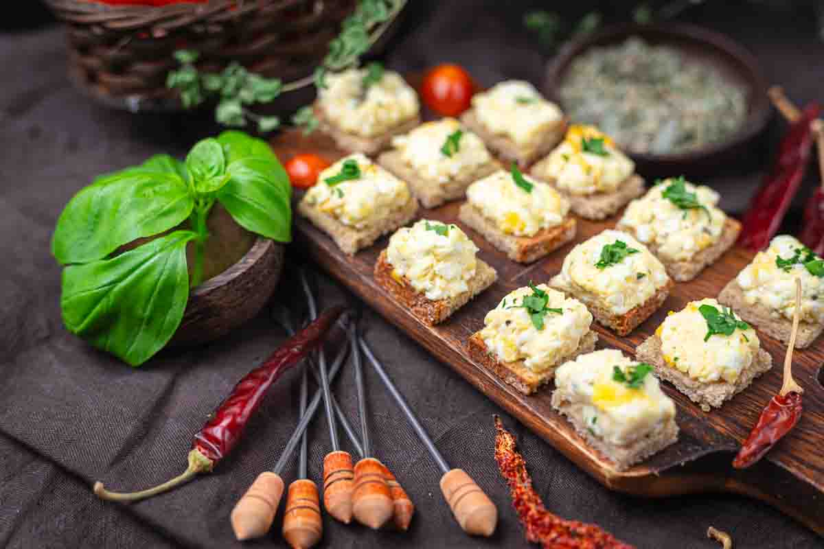 A wooden board displays several pieces of bread topped with a creamy mixture and herbs, surrounded by fresh basil, tomatoes, dried chili peppers, and skewers.