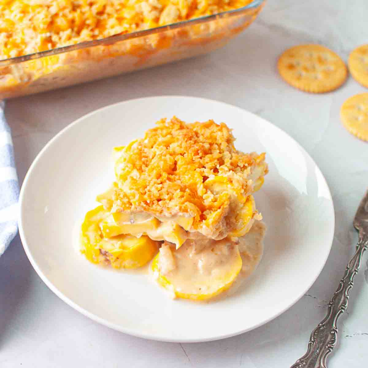 A serving of yellow squash casserole with a crumb topping on a white plate, with more casserole and round crackers in the background.