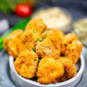 A bowl filled with golden-brown breaded and fried mushrooms, garnished with chopped herbs, with dipping sauces and vegetables in the background.