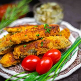 A plate of breaded, fried fish fillets garnished with fresh chives and cherry tomatoes, with sauces and herbs in the background.