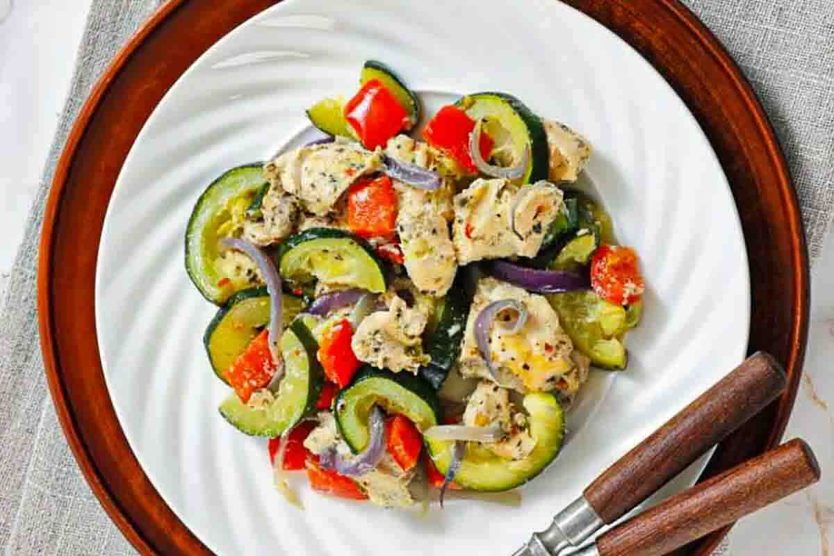 A plate of cooked chicken pieces mixed with zucchini, red bell peppers, and red onion slices, served on a white dish with wooden-handled utensils.