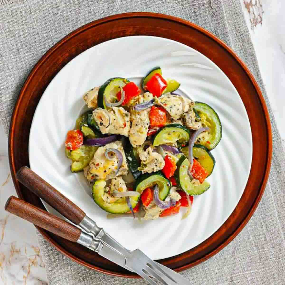 A white plate with a serving of baked chicken, zucchini, red bell pepper, and onions, set on a wooden charger. A fork and knife rest on the plate's edge.