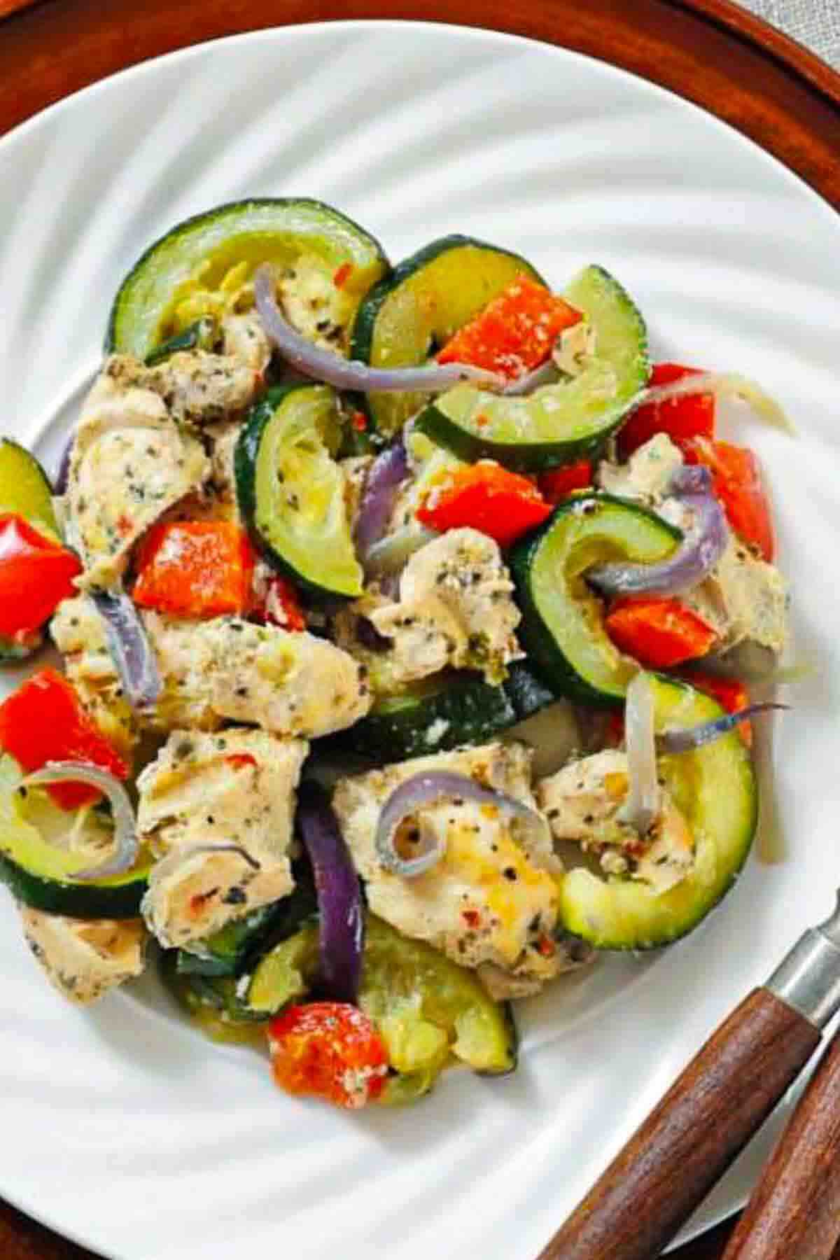 A white plate with a cooked dish of sliced zucchini, red bell pepper, red onion, and pieces of seasoned chicken. A fork and knife are placed to the right of the plate.