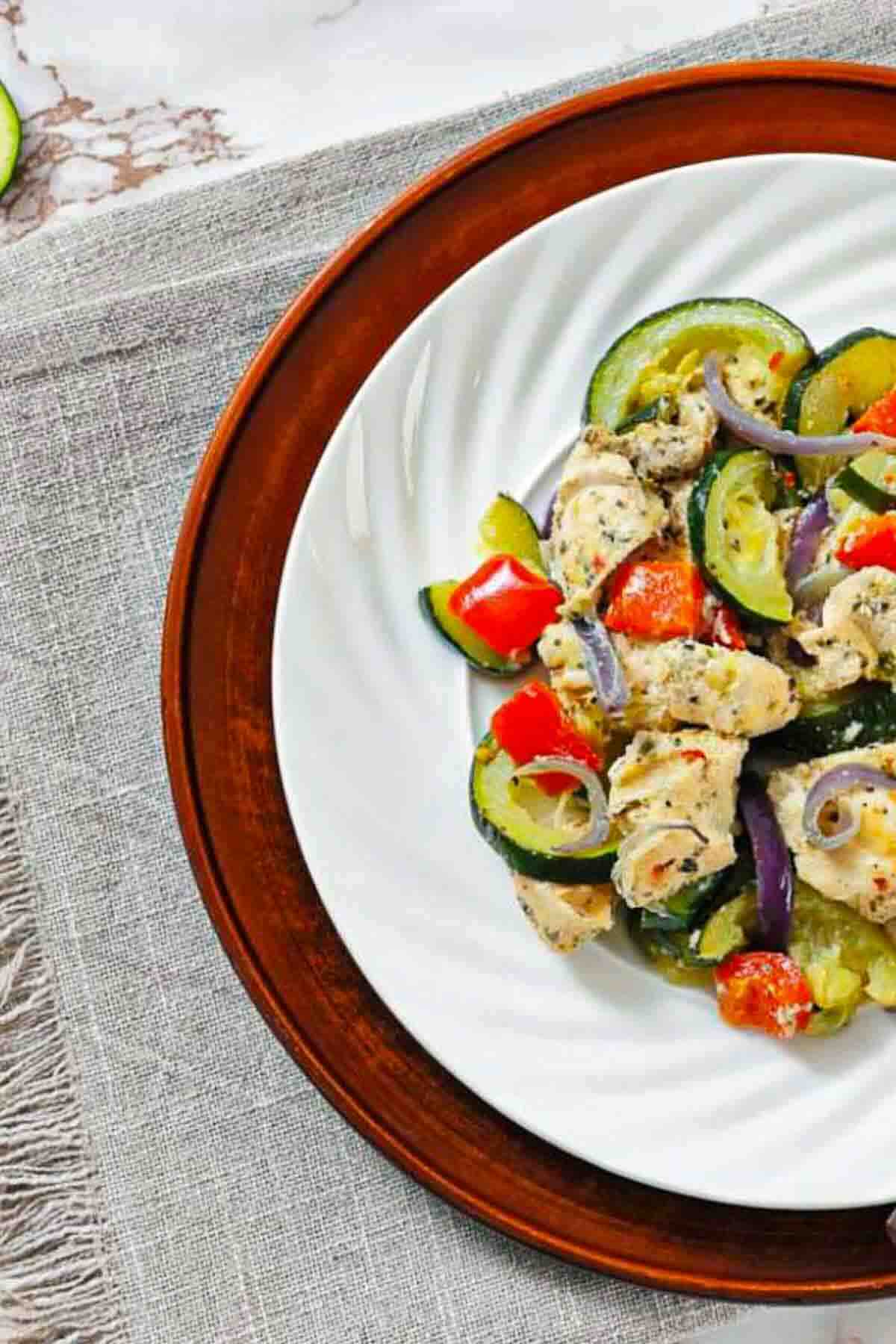A white plate with cooked chicken pieces, zucchini, red bell peppers, tomatoes, and red onions on a brown charger atop a gray cloth.