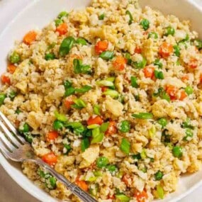 A bowl of cauliflower fried rice with peas, carrots, scrambled egg, and chopped green onions, served with a fork.