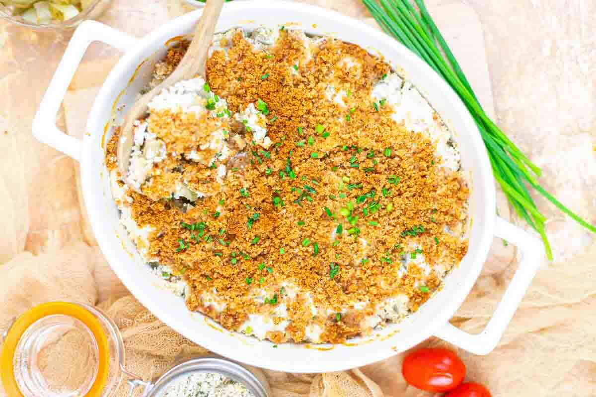 Casserole dish with tangy fried pickle ranch dip, placed on top of a brown cloth surrounded with tomatoes and chives.