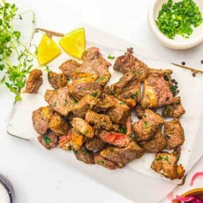 A serving tray with grilled steak bites garnished with herbs, lemon wedges, peppercorns, and parsley on a white background.