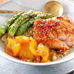 A bowl with grilled chicken, yellow bell peppers, asparagus spears, and quinoa, garnished with sesame seeds and red chili threads, served with a spoon.