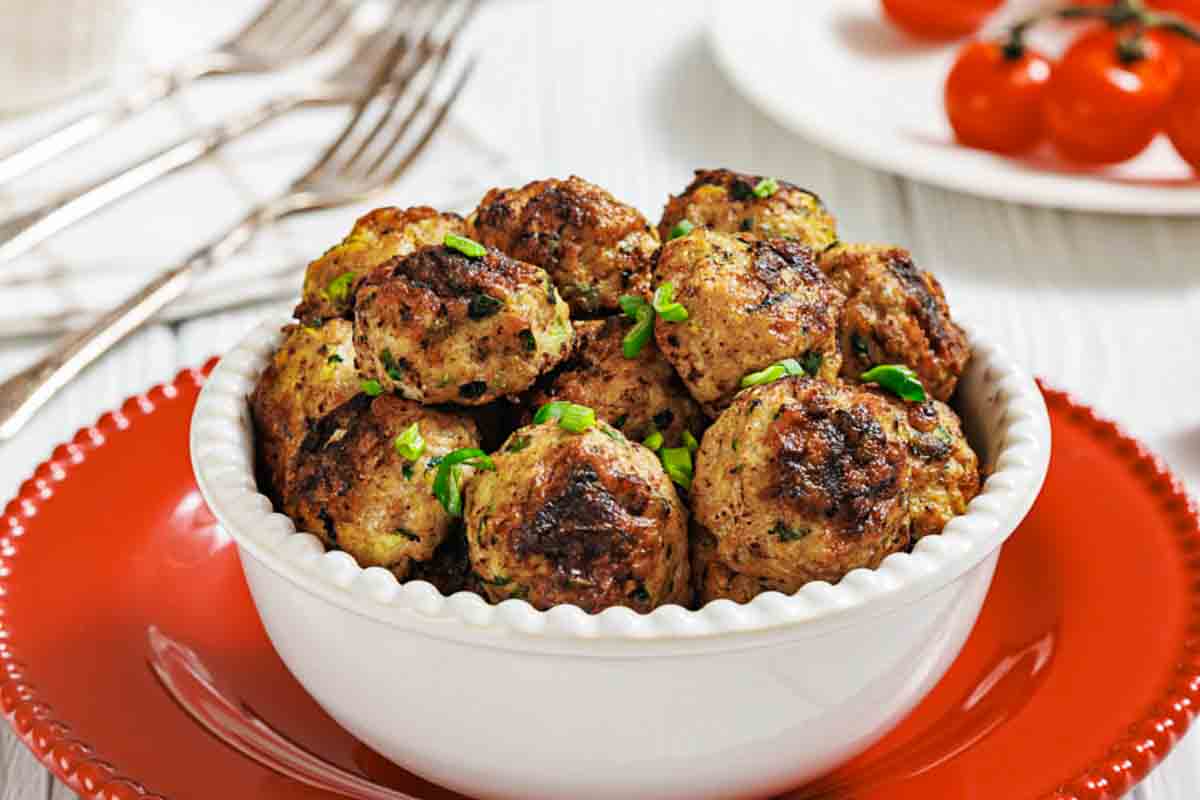 A white bowl filled with browned chicken poppers garnished with chopped herbs sits on a red plate; tomatoes and forks are in the background.