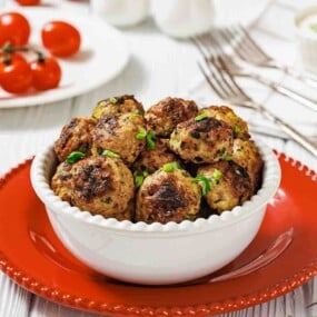 A white bowl filled with cooked meatballs garnished with herbs, placed on a red plate, with tomatoes and cutlery in the background.