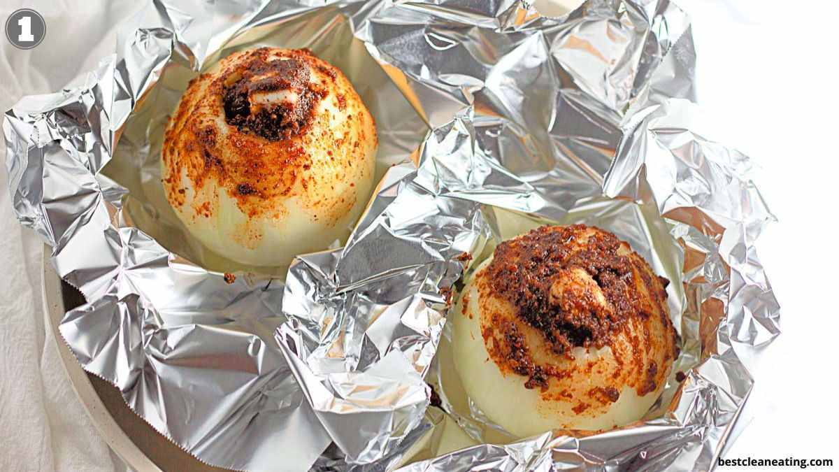 Two whole onions topped with seasoning and butter sit on aluminum foil, ready to be baked.