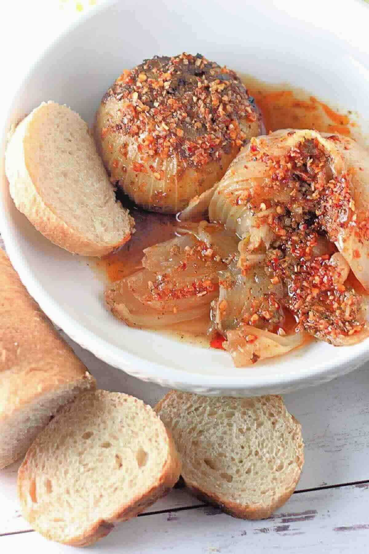 A white bowl with roasted onion topped with spices and oil, accompanied by sliced bread on the side.