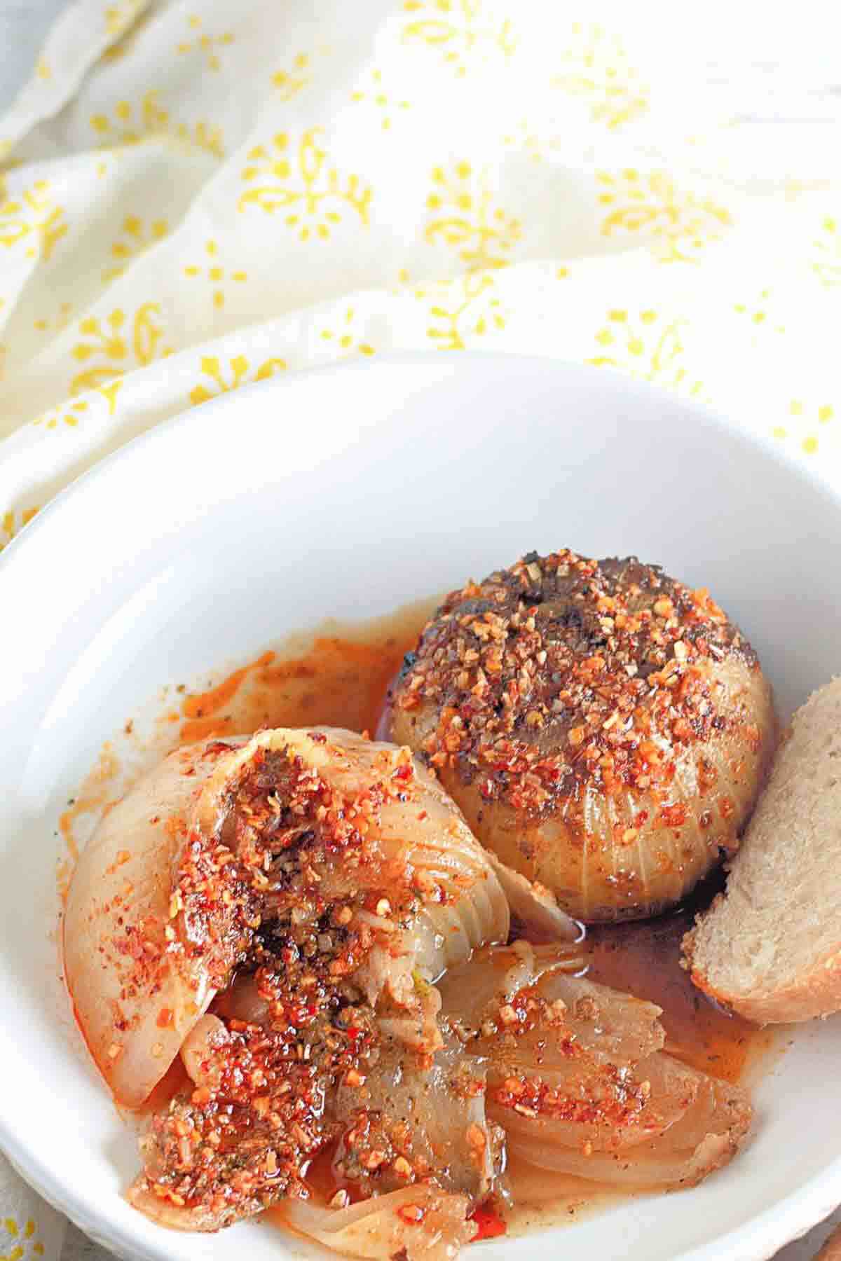 A white bowl contains two baked stuffed onions topped with spices and breadcrumbs, sitting in a light sauce, with a slice of bread on the side.