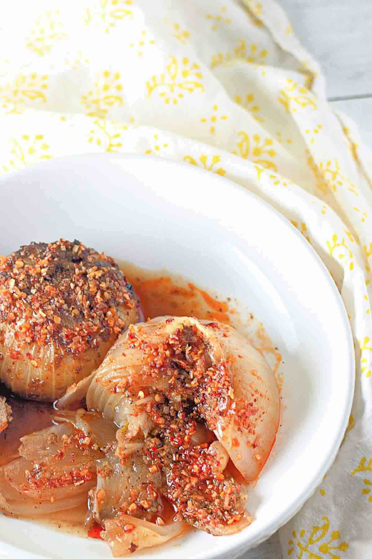 A white bowl containing stuffed onions topped with a red spice mixture, placed on a light surface with a yellow-patterned cloth in the background.
