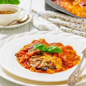 A plate of baked eggplant parmesan topped with tomato sauce and fresh basil, served with a fork on a white plate. A pan of the dish and a bowl of spices are in the background.