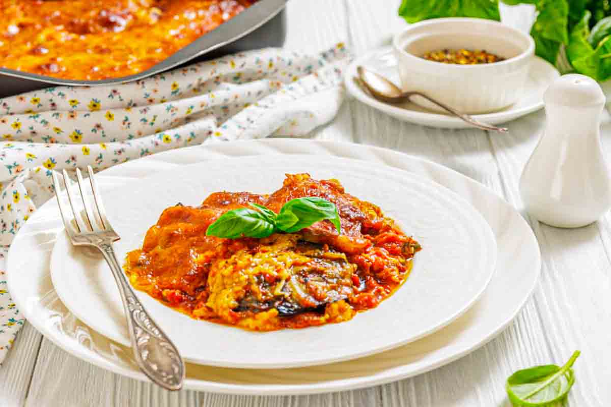 A serving of baked eggplant parmesan on a white plate, garnished with fresh basil, with a fork and knife beside it on a light table setting.