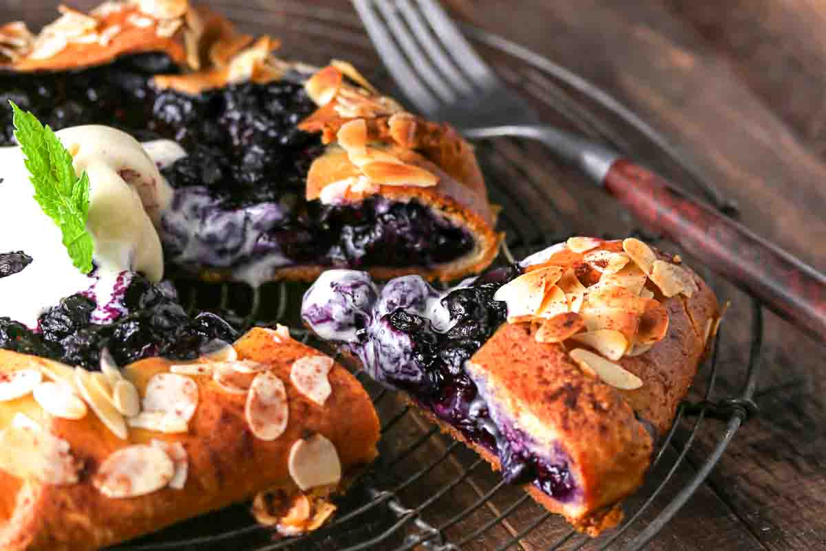 A sliced blueberry galette topped with almond flakes and a scoop of vanilla ice cream, served on a wire cooling rack with a fork nearby.