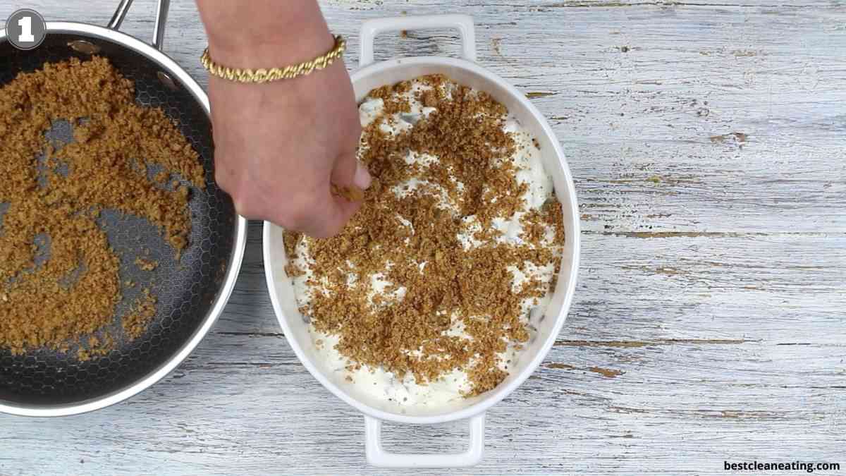 A hand sprinkles brown crumb topping onto a white dish filled with a creamy mixture, with extra crumbs in a black pan nearby, on a light wooden surface.