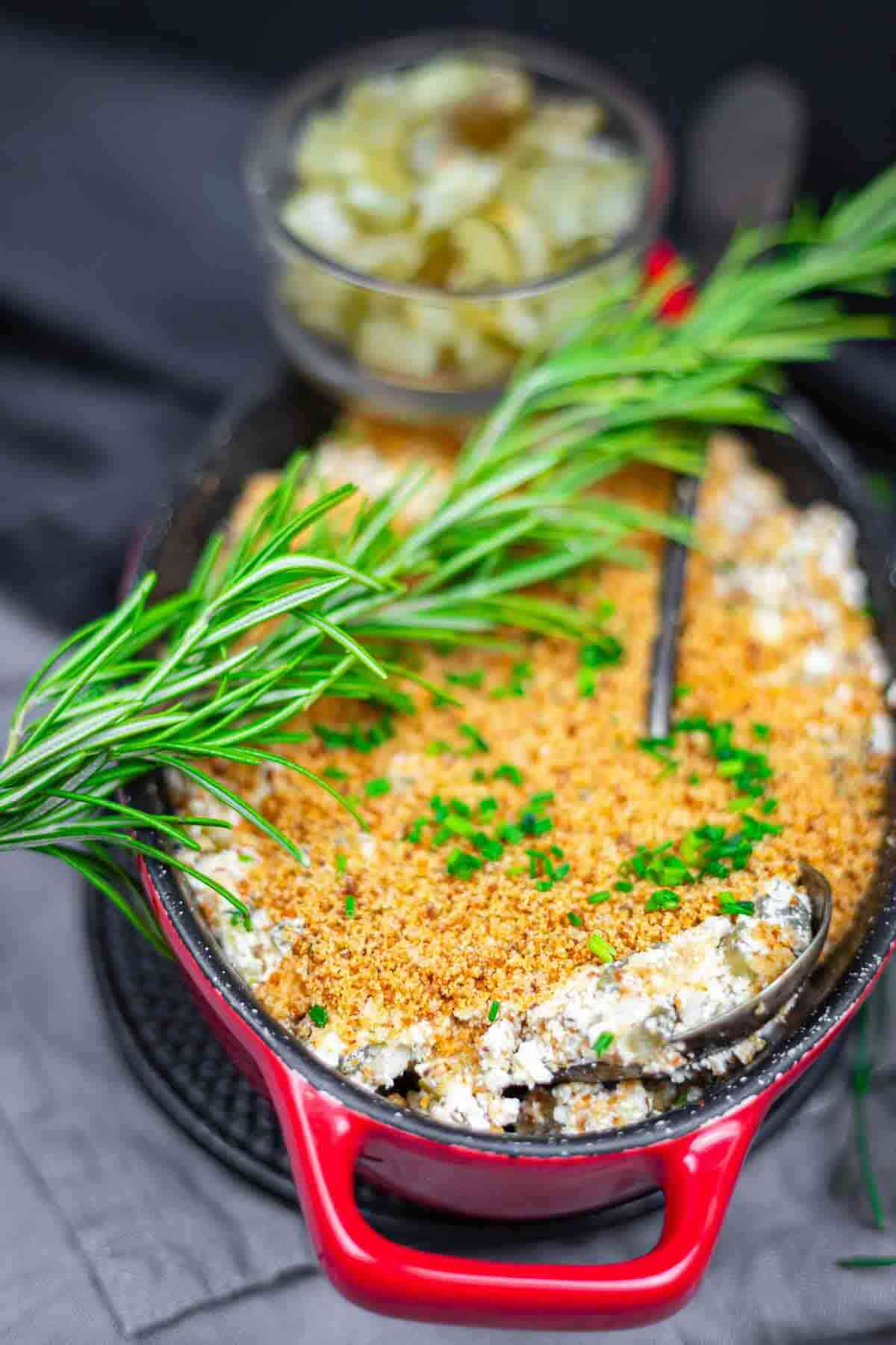 A red baking dish filled with a breadcrumb-topped casserole, garnished with fresh rosemary and chopped chives, with a serving spoon and a bowl of grapes in the background.
