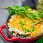 A red baking dish filled with a breadcrumb-topped casserole, garnished with fresh rosemary and chopped herbs, with serving spoons inside.