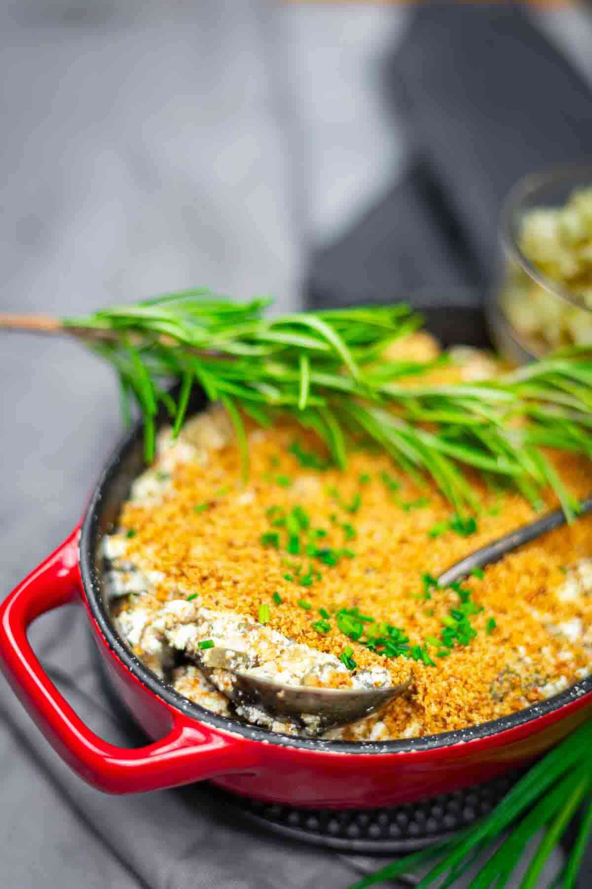 A red casserole dish filled with a breadcrumb-topped baked dish, garnished with fresh herbs and a sprig of rosemary, with a spoon inside.