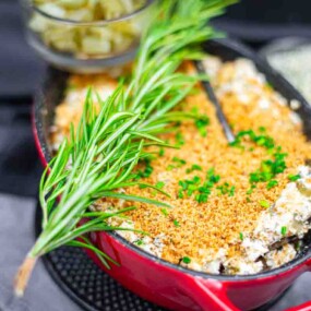 A baked casserole topped with golden breadcrumbs and chopped herbs is served in a red dish, garnished with a sprig of rosemary.