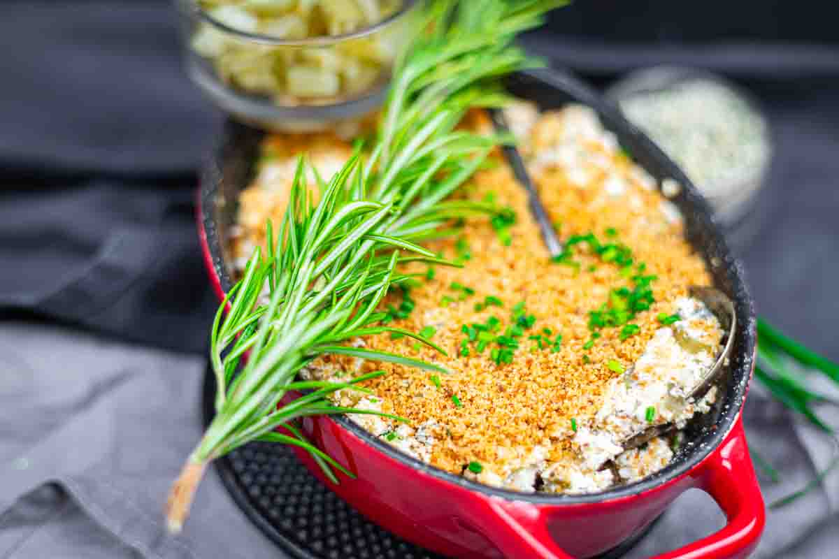 A baked casserole topped with golden breadcrumbs and chopped herbs is served in a red dish, garnished with a sprig of rosemary.