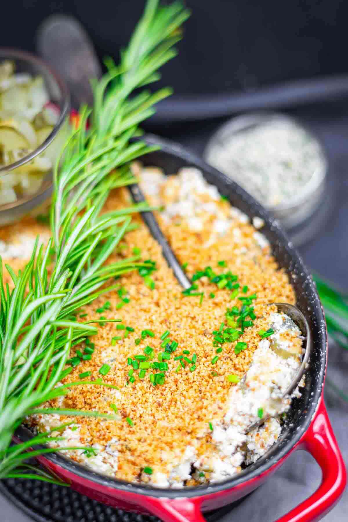 A casserole dish filled with a breadcrumb-topped baked dip, garnished with chopped herbs, with fresh rosemary placed on the side and a metal serving spoon inside.