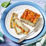 A slice of baked egg casserole with spinach and cheese is served on a white plate with a fork, beside two thin cut pieces.