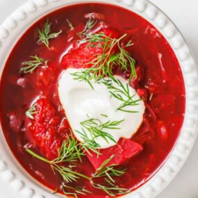 A bowl of red beet soup topped with a dollop of sour cream and fresh dill, served on a white plate.