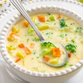 A bowl of creamy vegetable soup with broccoli, carrots, and potatoes, with a spoon inside the bowl on a white plate.