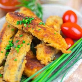 A plate of breaded and fried fish sticks garnished with chopped chives, served with fresh cherry tomatoes and herbs.