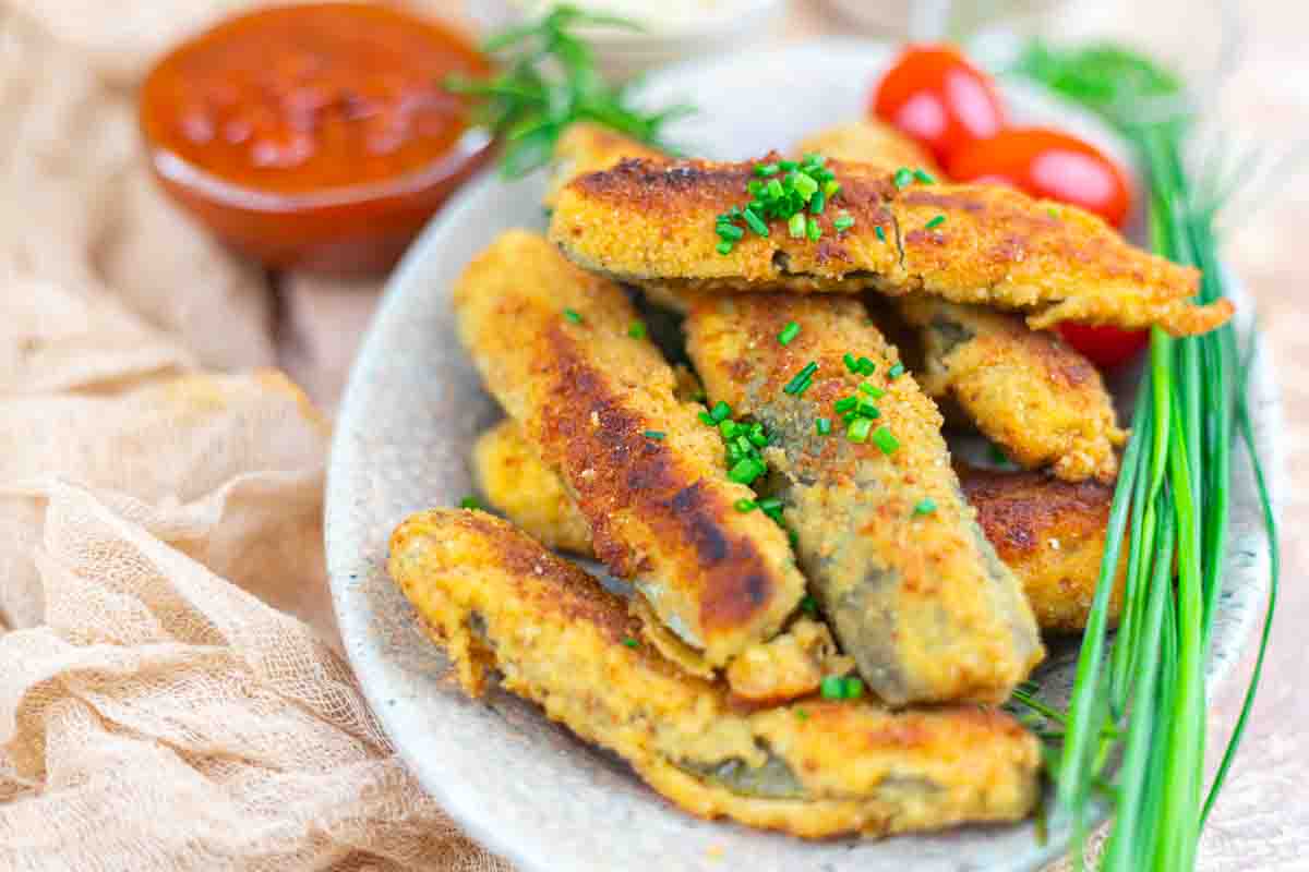 A plate of breaded and fried pickle sticks garnished with chopped chives, served with cherry tomatoes and a bowl of red dipping sauce.