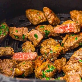 Cubed pieces of steak, seasoned with herbs and slightly browned, sit in a black air fryer basket.