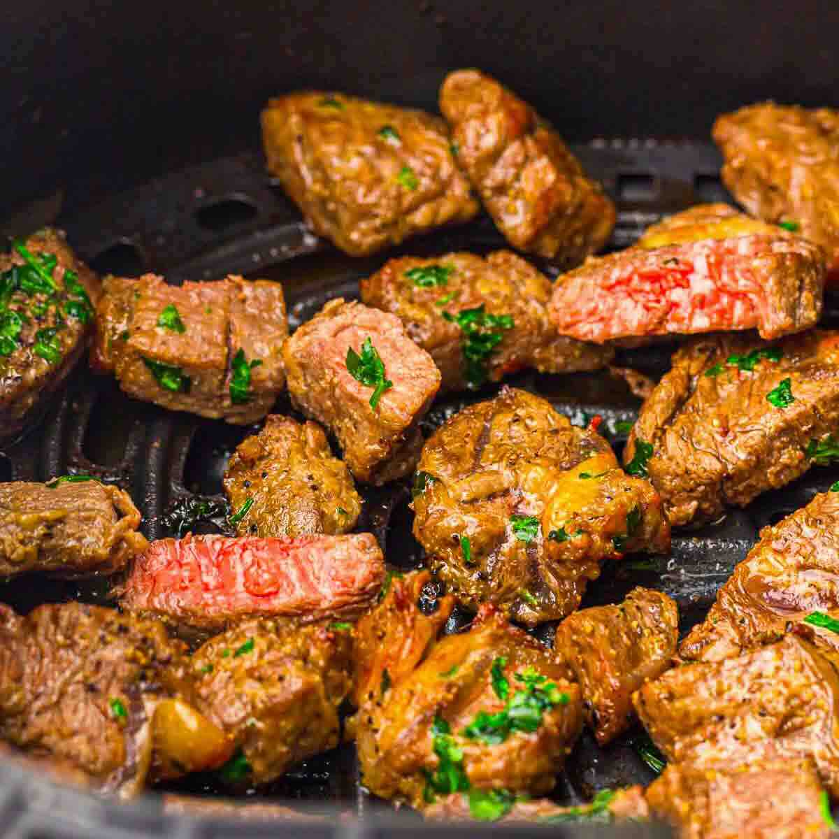 Cubed pieces of steak, seasoned with herbs and slightly browned, sit in a black air fryer basket.