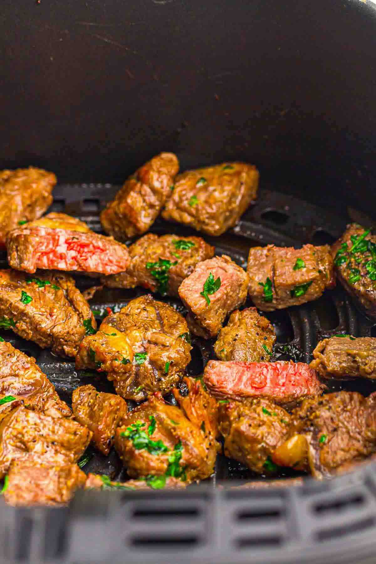 Chunks of cooked steak with herbs are shown inside an air fryer basket.