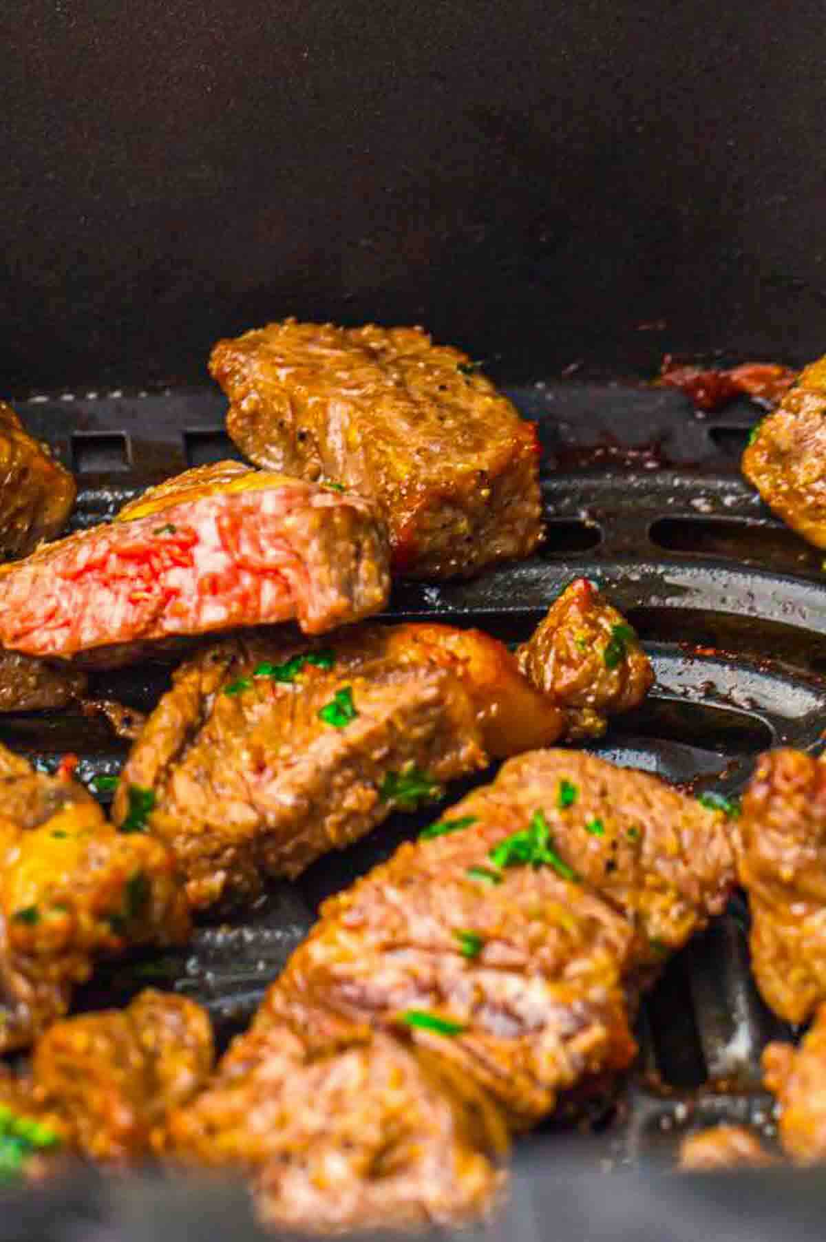 Several pieces of cooked steak bites, some showing a pink center, on a black grill or air fryer tray.