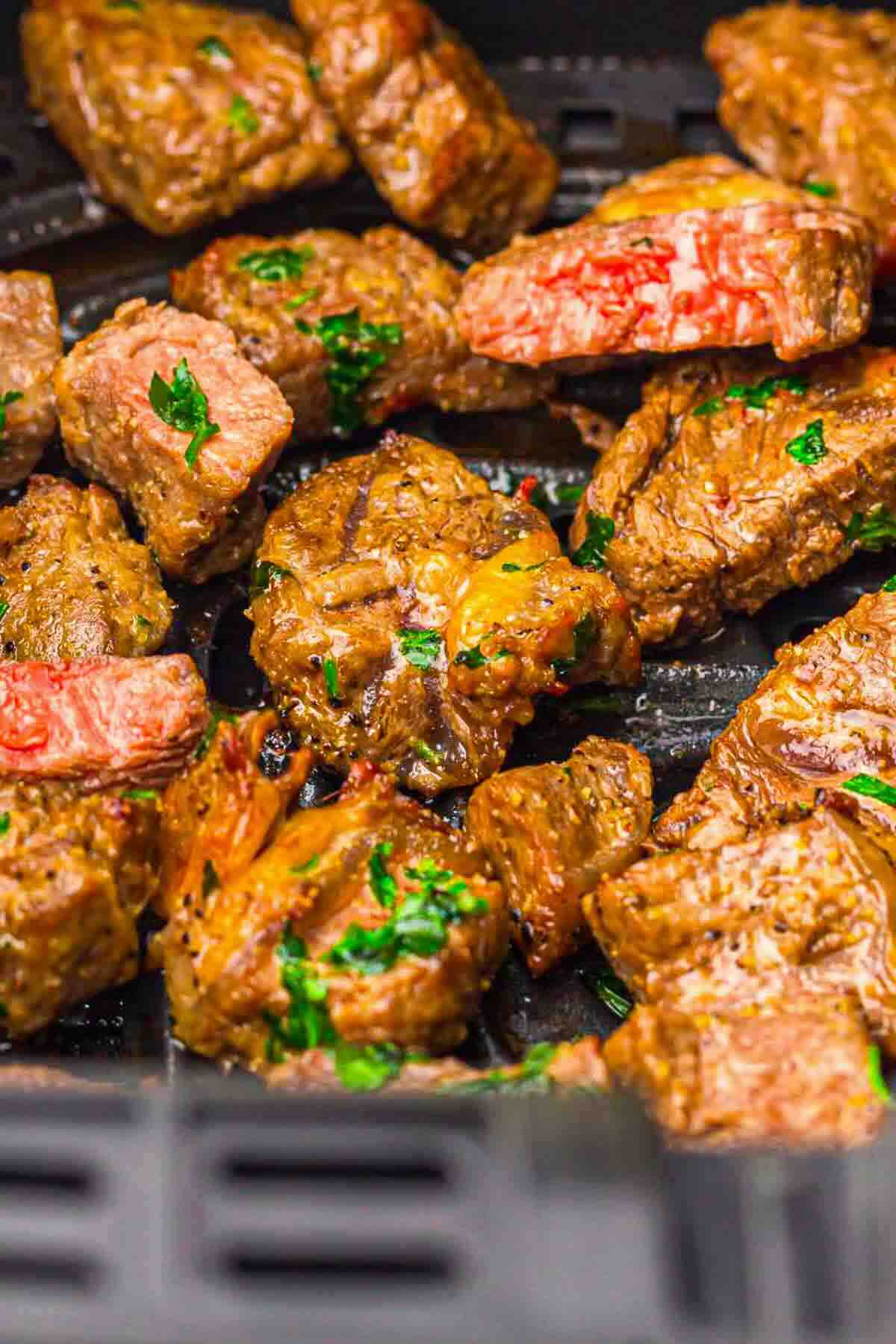 Cooked steak bites with herbs are arranged in an air fryer basket.